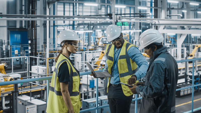 Young Diverse Team of Car Factory Engineers in High Visibility Vests Using Laptop Computer. Automotive Industrial Manufacturing Facility Working on Vehicle Production. Diversity on Assembly Plant.