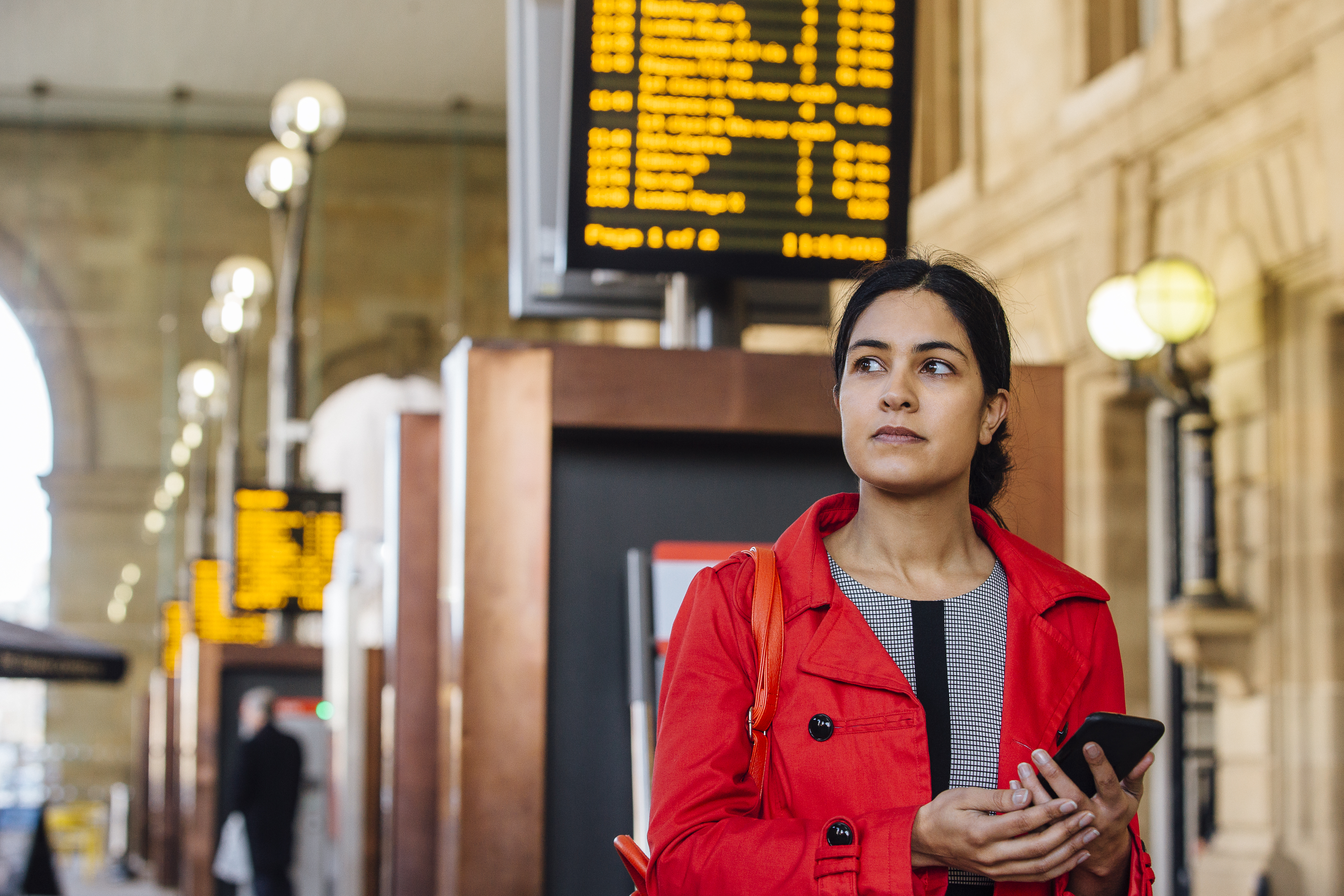 Voyageuse au premier plan tenant un smartphone dans une gare avec des panneaux d'affichage électroniques.
