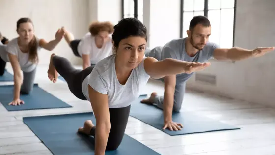 Concentrated young female indian trainer showing bird dog pose at group yoga class. Fit barefoot diverse people practicing body balancing exercise in Parsva Balasana position on mat indoors in gym.
