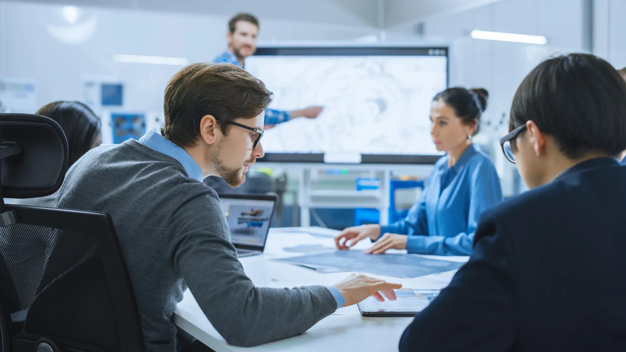 Salle de réunion moderne des bureaux d’usine : une équipe diversifiée d’ingénieurs, de gestionnaires et d’investisseurs qui discutent à la table de conférence, utilisent la télévision interactive, analysent, trouvent des solutions, discutent des concepts.