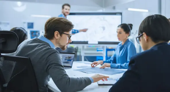 Ein moderner Besprechungsraum in einer Fabrik: Ein vielbeschäftigtes und vielfältiges Team aus Ingenieuren, Managern und Investoren sitzt an einem Konferenztisch, nutzt interaktives Fernsehen, analysiert, erarbeitet Lösungen und diskutiert Konzepte.