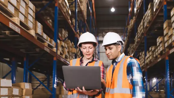 two warehouse workers looking at a laptop together in a warehouse facility wearing ppe (hard hats and orange vests)
