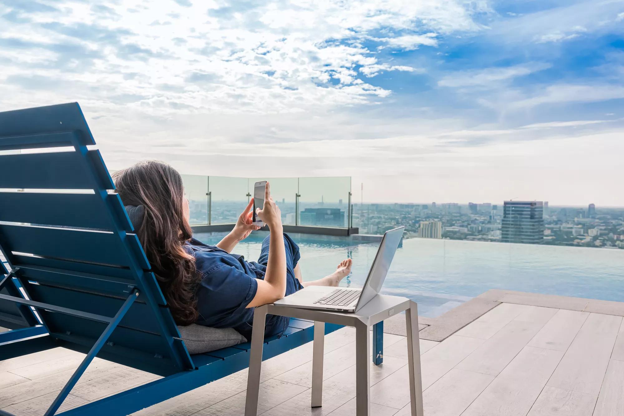 Summer day lifestyle asian woman relax and chill near luxury swimming pool. Asian woman using app on a smartphone and working on a laptop and Blurred background skyscraper, Beautiful sky and clouds.