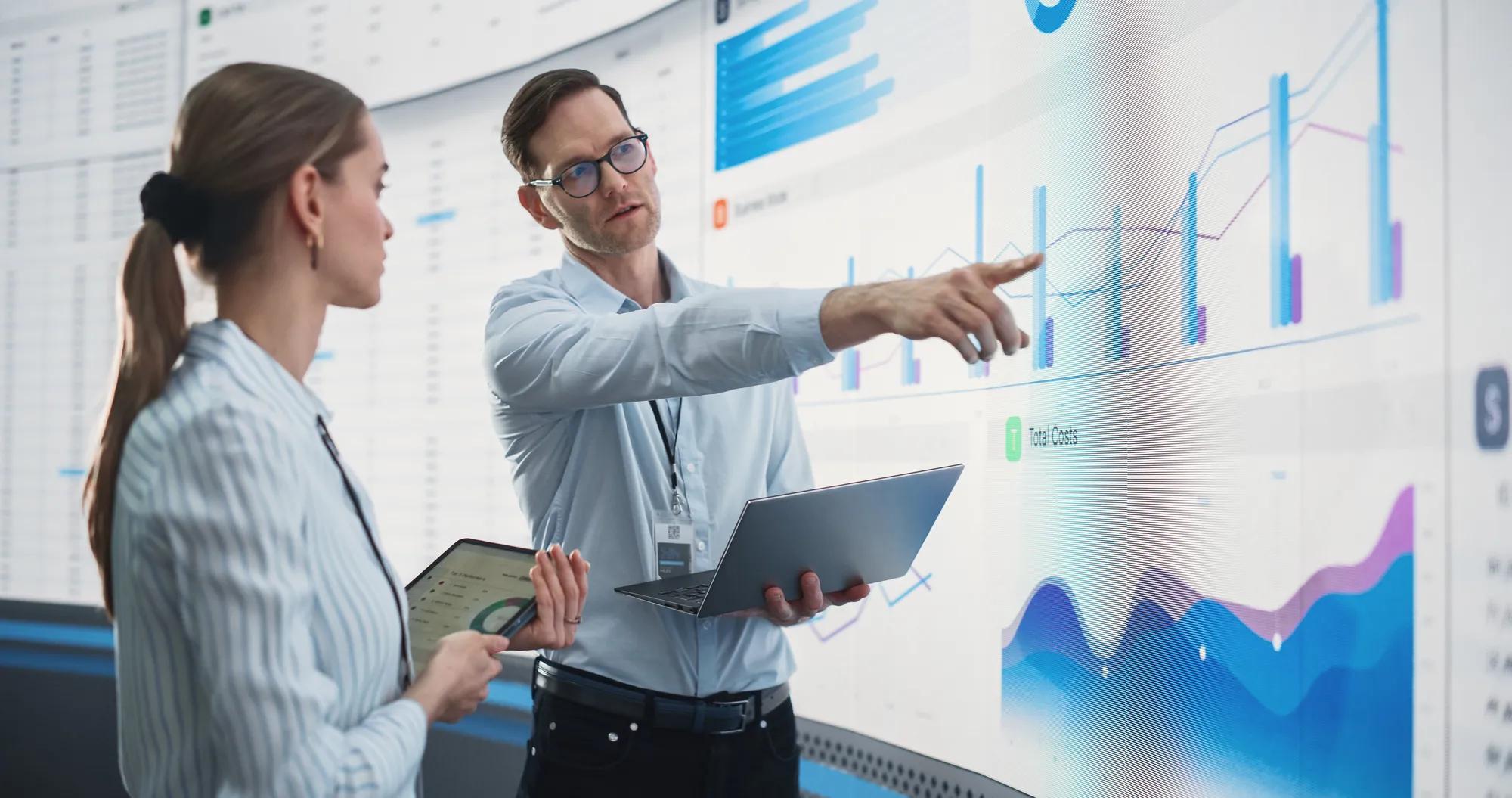 Caucasian Male And Female Data Scientists With Laptop And Tablet Standing Next To Big Digital Screen With Graphs And Charts In Monitoring Office. Colleagues Discussing Business Opportunities Or Risks.