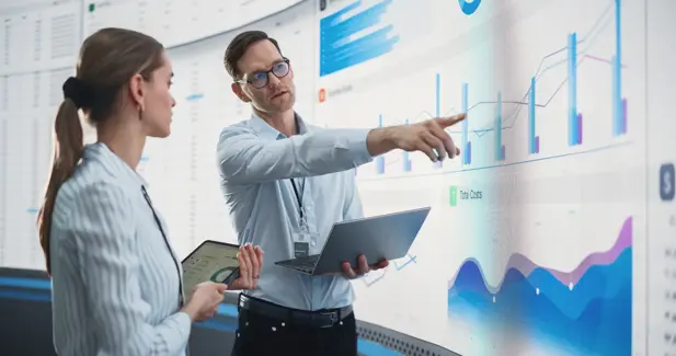 caucasian male and female data scientists with laptop and tablet standing next to big digital screen with graphs and charts in monitoring office. colleagues discussing business opportunities or risks.