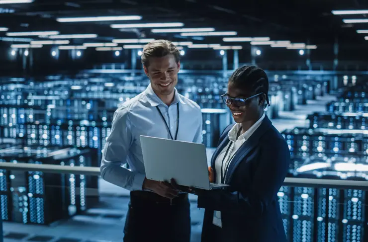 Eine Systemadministratorin und ein IT-Spezialist unterhalten sich im Rechenzentrum und benutzen dabei einen Laptop. Informationstechnologie-Ingenieure arbeiten an der Cybersicherheitsabsicherung in Cloud-Computing-Serverfarmen.