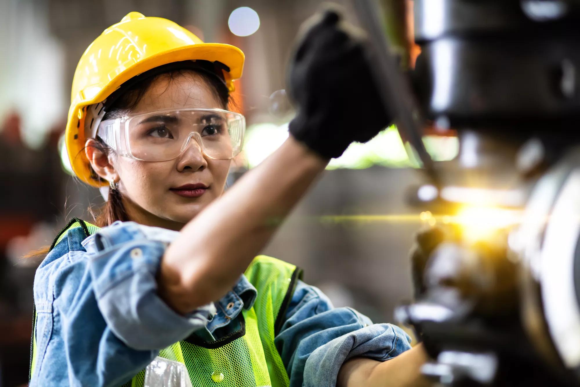 Woman worker wearing safety goggles control lathe machine to drill components. Metal lathe industrial manufacturing factory