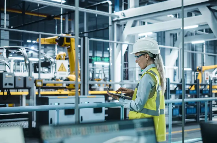 Usine automobile : ingénieure automobile portant un casque de sécurité, debout, utilisant un ordinateur portable. Surveillance, contrôle, production d’équipements. Chaîne de montage automatisée de bras robotisés fabriquant des véhicules électriques.