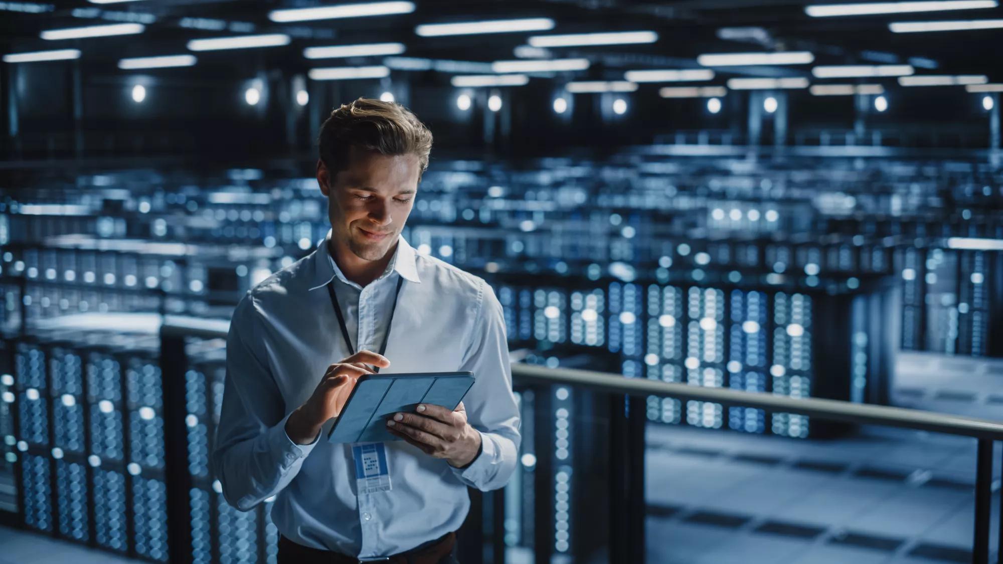 Portrait of Smiling IT Specialist Using Tablet Computer in Data Center. Big Server Farm Cloud Computing Facility with Male Maintenance Administrator Working. Cyber Security, e-Business.