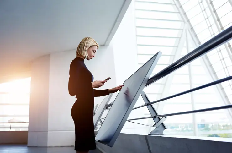 Female architect holding cell telephone while viewing 3d-visualization of project on digital touch screen, woman preparing a presentation for staff via high tech computer display and mobile phone