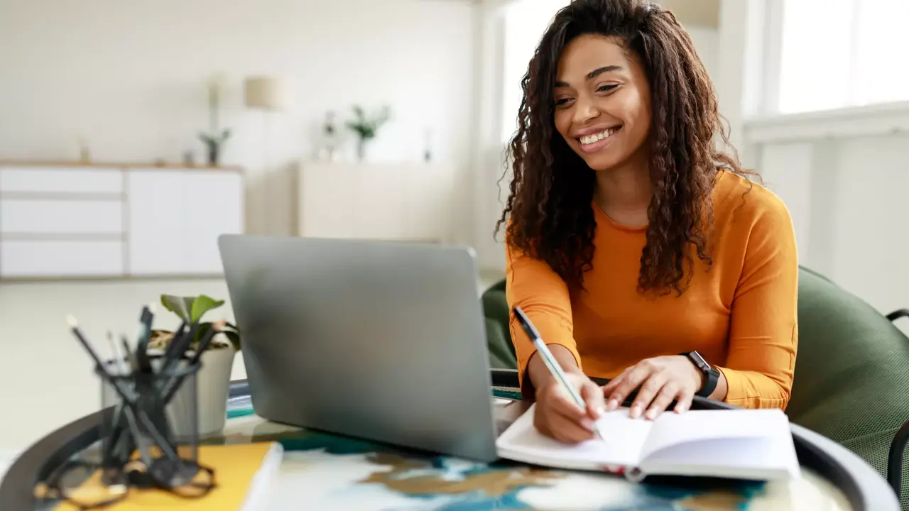 smiling young african american woman sitting at desk working on laptop taking notes in notebook, happy millennial female studying online, watching webinar using computer and writing check list