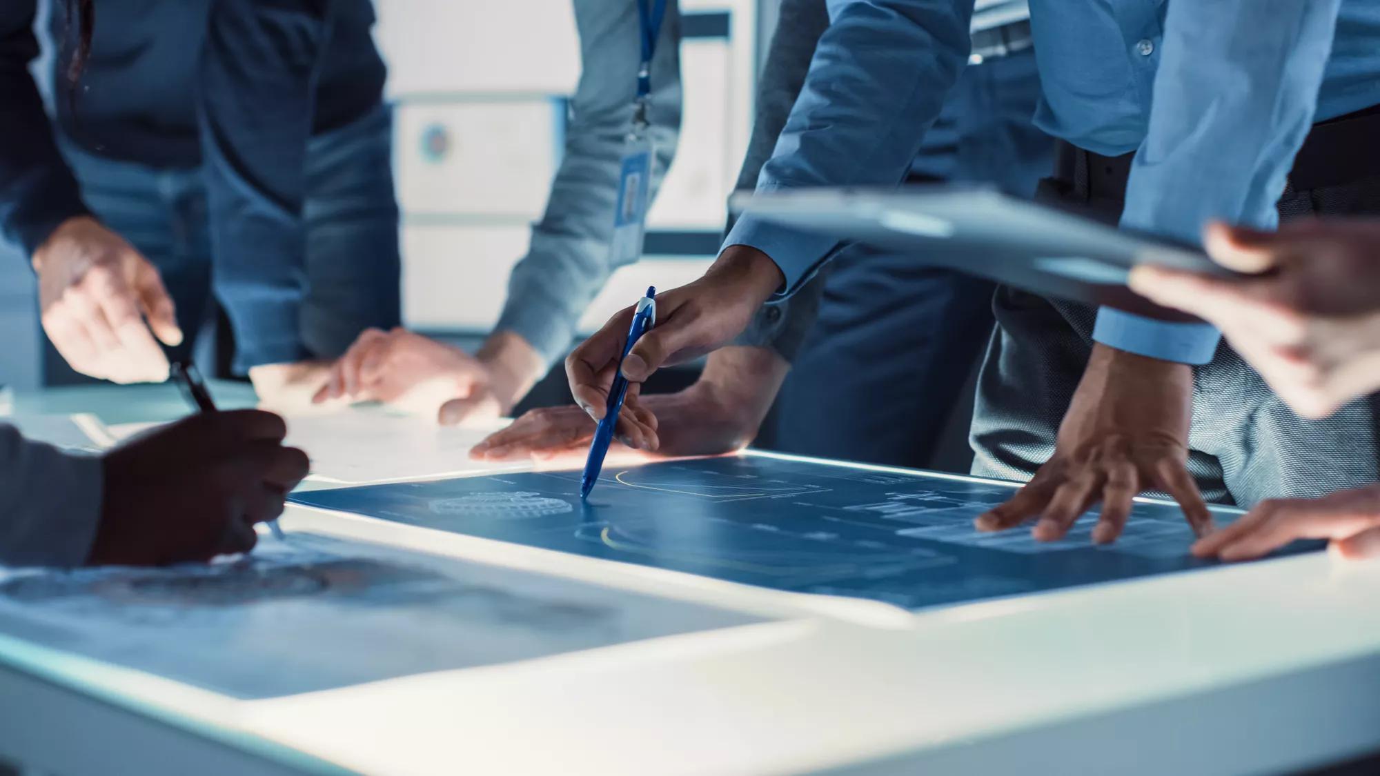 Des ingénieurs, des scientifiques et des développeurs réunis autour d’une table de conférence illuminée dans le centre de recherche technologique, discutant, trouvant des solutions et analysant la conception de moteurs industriels. Plan de mains rapprochées