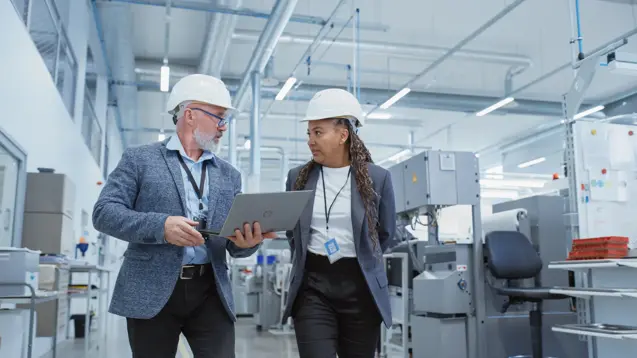 portrait of a two diverse heavy industry engineers in hard hats walking with laptop computer and talking in a factory. footage of two manufacturing employees at work.