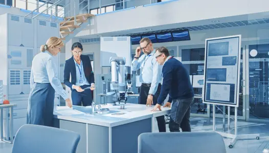 Diverse Team of Industrial Robotics Engineers Gathered Around Table With Robot Arm, They Manipulate and Program it to Pick Up and Move Metal Component. Bright Facility, Creative People