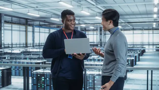 Two Multiethnic Male Data Center System Administrators and IT Specialists Talk, Use Laptop. Information Technology Engineers work on Cyber Security Network Protection in Cloud Computing Server Farm.