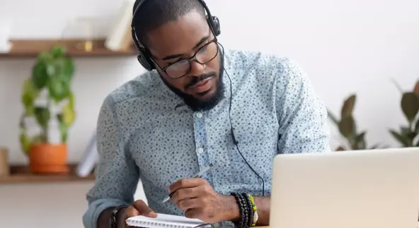 focused african business man in headphones writing notes in notebook watching webinar video course, serious black male student looking at laptop listening lecture study online on computer e learning