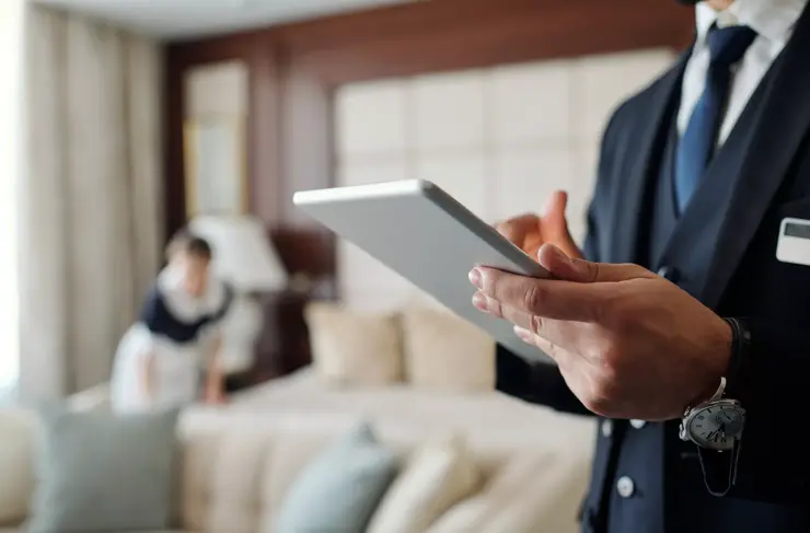 Nahaufnahme eines jungen männlichen Hotelmanagers mit digitalem Tablet, während er die Liste der gebuchten Gäste mit Zimmermädchen im Hintergrund durchsieht