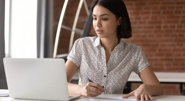 Focused asian business woman working studying online in office looking at laptop making notes, serious japanese employee or student watching webinar writing information in notebook sitting at desk